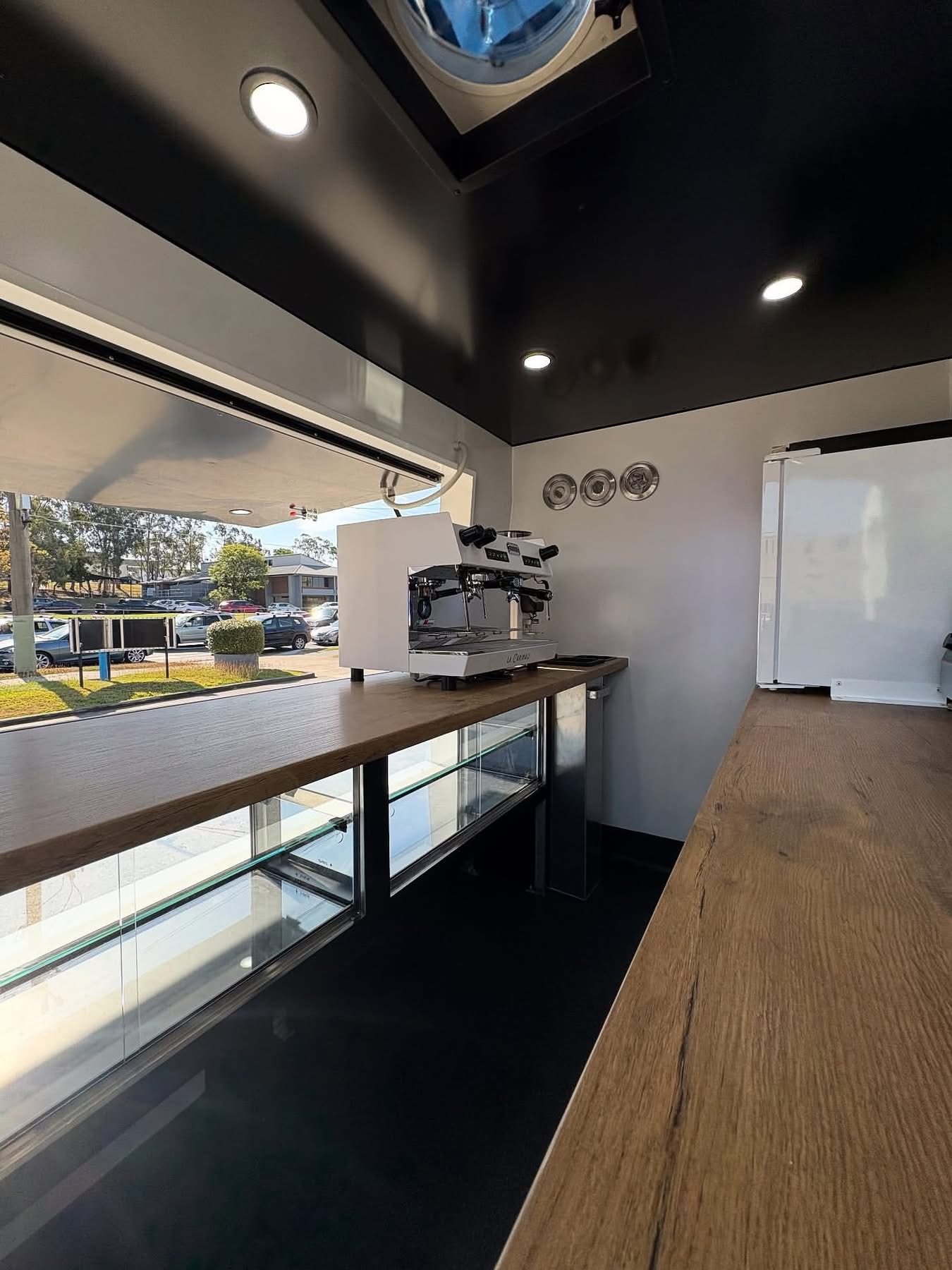 Interior of a walk-in coffee van with timber benches, a white commercial espresso machine and a glass display cabinet